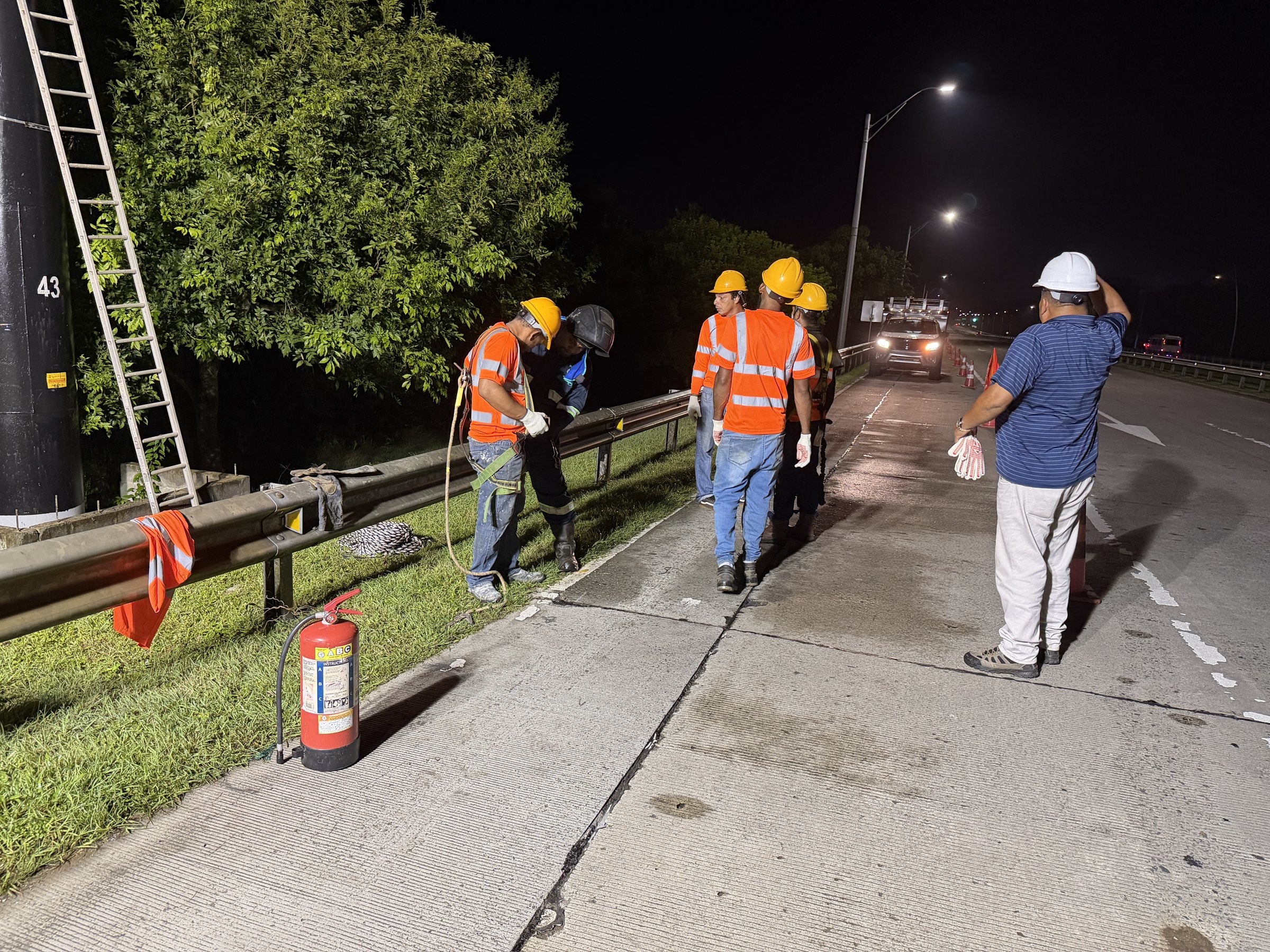 Nighttime safety briefing with crew members in protective gear beside a highway unipole